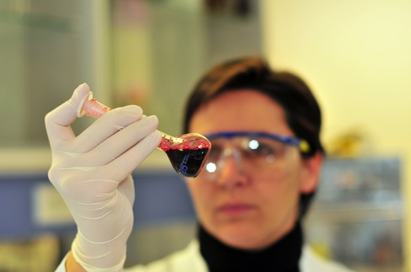 Scientist wearing protective eyewear and gloves examining a vial containing a dark red liquid in a laboratory setting.