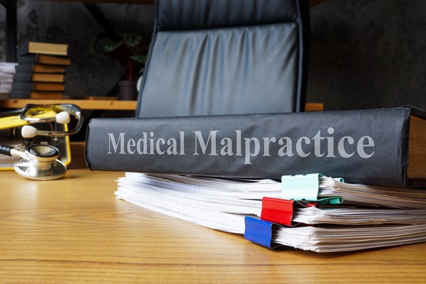 Large binder labeled “Medical Malpractice” resting on a stack of clipped documents on a wooden desk beside a stethoscope and an office chair.