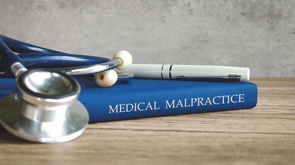 A blue book with the words "MEDICAL MALPRACTICE" printed on the spine, lying on a wooden surface with a stethoscope and pen resting on top